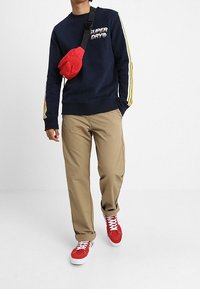 Persona vistiendo un sudadero azul marino con rayas amarillas, pantalones caqui, una bolsa cruzada roja y zapatillas rojas caminando frente a un fondo blanco.