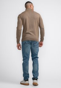 Beige gestrickter Pullover mit hohem Kragen, kombiniert mit blauen Jeans. Das Outfit zeichnet sich durch figurbetonte Ärmel und eine strukturierte Oberfläche aus.