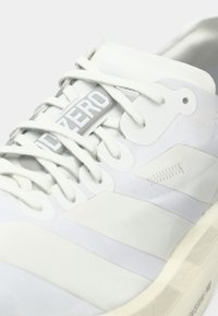 Gros plan sur une sneaker blanche avec des lacets, marquée "DOZER0" sur la languette, et un motif cousu sur le panneau latéral.