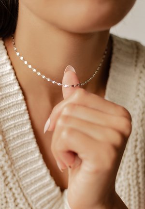 Femme portant un pull en maille beige tient un collier en chaîne argentée en forme de cœur près de son cou entre le pouce et l'index.