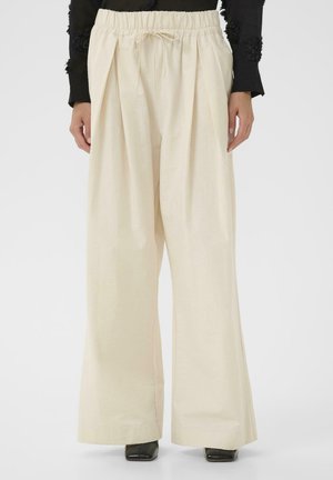 Krémové široké nohavice s vysokým pásom, so sťahovacou elastickou páskou a šnúrkou, nosené s čiernym topom a čiernymi topánkami so štvorcovou špičkou.