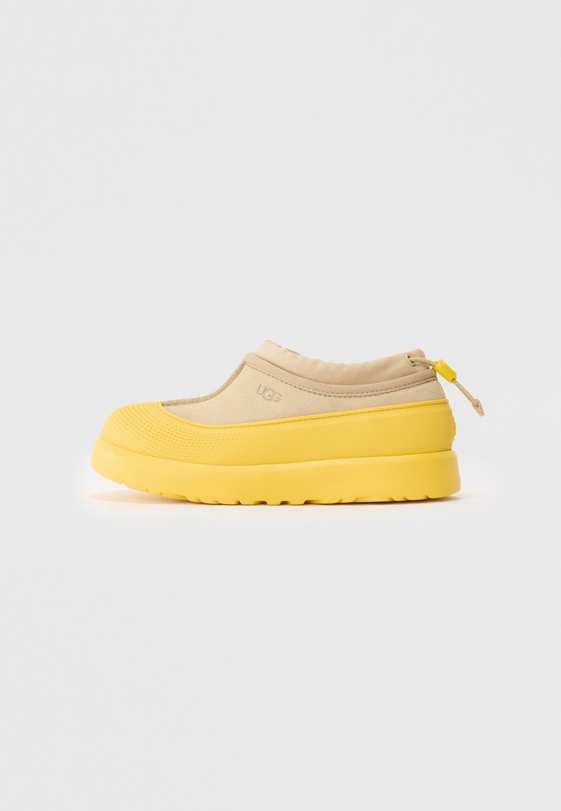 Chaussure beige à enfiler avec une semelle épaisse et texturée jaune, un bout renforcé, un col de cheville élastique et une languette arrière avec un bouton jaune.