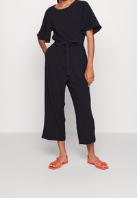 Mono negro con mangas cortas, ajuste holgado y cintura con lazo. Combinado con sandalias de tiras de color naranja brillante. Textura suave, diseño minimalista.