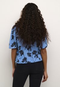 Blaue Bluse mit lockerer Passform, kurzen Ärmeln, die mit schwarzen Blumenmustern verziert ist, getragen mit schwarzen Jeans.
