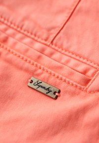 Tessuto corallo con una texture morbida, caratterizzato da cuciture orizzontali e un'etichetta metallica con la scritta "Superdry" attaccata.