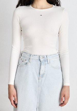 Woman wearing a fitted long-sleeve white ribbed top and a high-waisted light blue denim skirt, standing with arms relaxed.