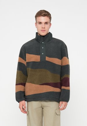 Fleece-pusero, jossa on korkea kaulus tummanharmaana, ja jossa on aaltoilevia raitoja ruskeassa, oliivinvihreässä ja burgundin värissä. Kolmen napin listatiedot.