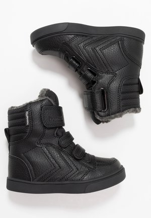 Paire de baskets montantes en cuir noir avec bandes velcro et doublure en fourrure grise à l'intérieur.