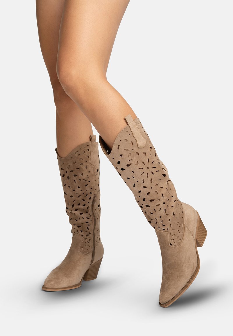 Frau trägt beige Wildlederstiefel bis zur Mitte der Wade mit floralen Ausschnitten, seitlichem Reißverschluss und gestapelten Holzabsätzen, hebt leicht einen Fuß an.