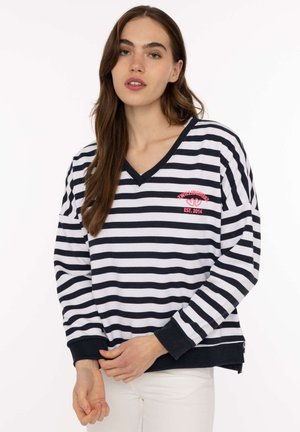 Frau trägt einen schwarz-weiß horizontal gestreiften V-Ausschnitt-Pullover mit umgeschlagenen Ärmeln und weiße Hose, steht vor einfarbigem Hintergrund.