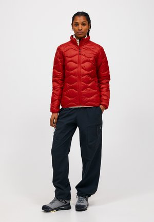 Femme debout portant une veste matelassée rouge, un pantalon cargo noir et des chaussures de randonnée grises sur un fond blanc uni.