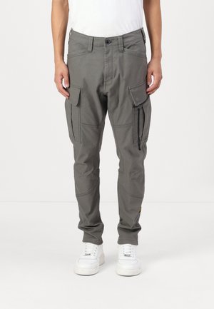 Mand iført grå cargobukser med klaplommer og hvide sneakers, der står mod en ensartet hvid baggrund.