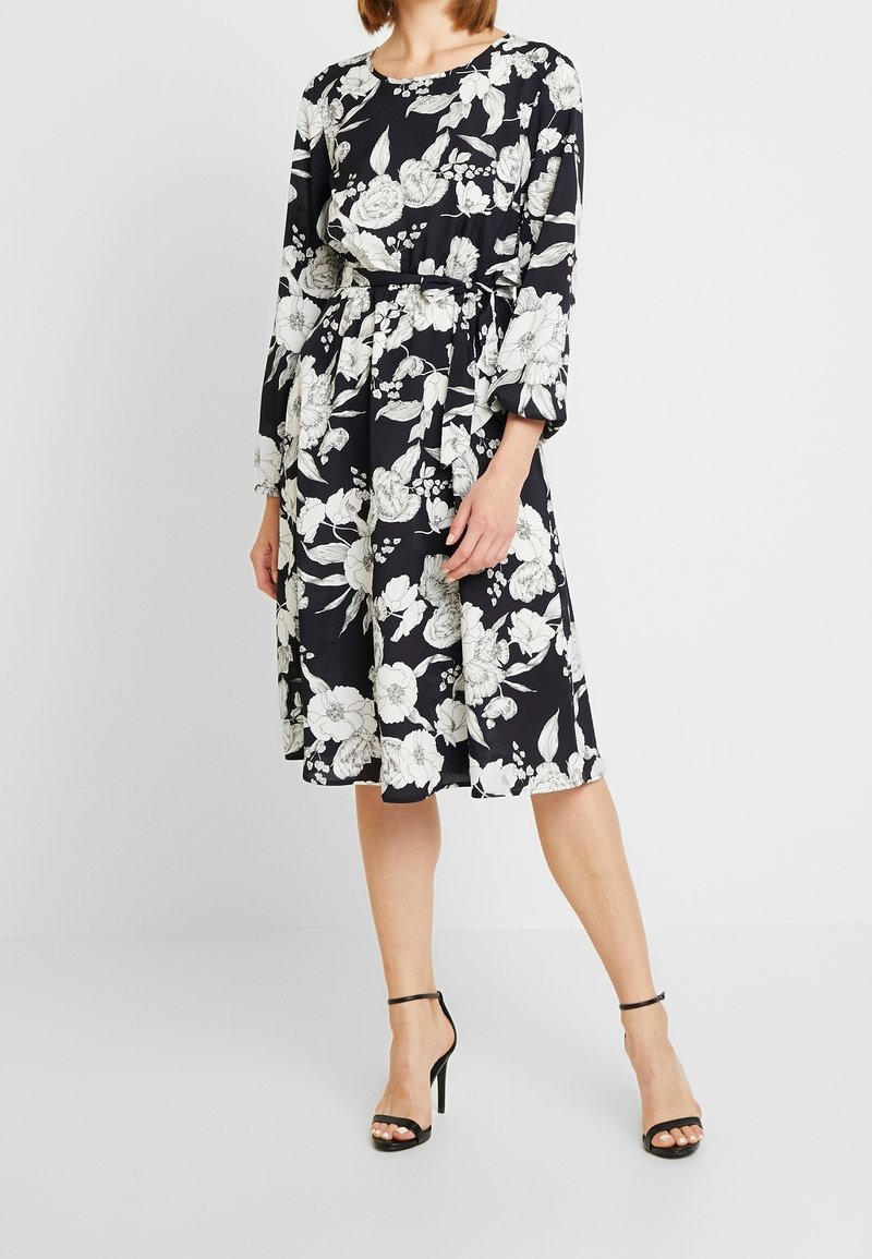 Floral dress in black with white flowers, long sleeves, a belted waist, and a knee-length skirt. Styled with black high-heeled sandals.
