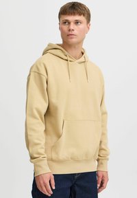 Sudadera con capucha beige en una tela suave, con un bolsillo tipo canguro, puños y dobladillo acanalados, con cordones y detalles de metal plateado.