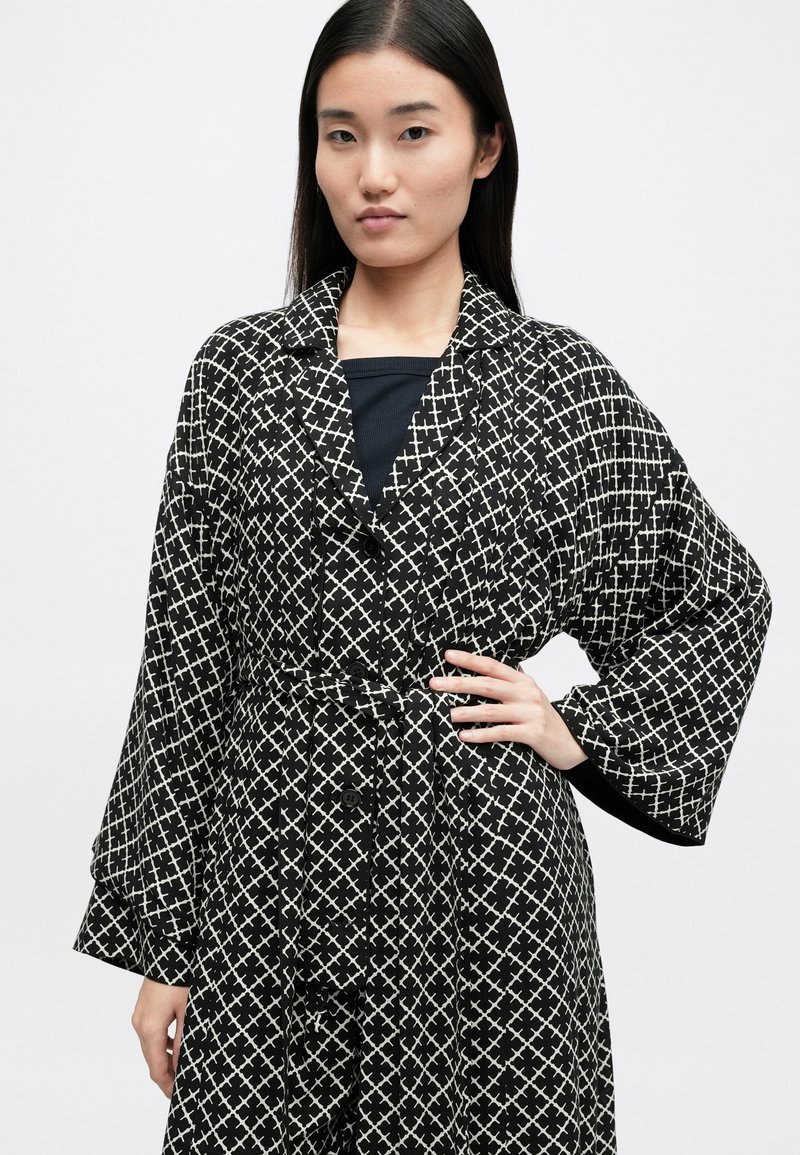 Schwarz-weiß gemustertes Gewand mit tailliertem Schnitt, großen Ärmeln und Knöpfen vorne. Der Stoff hat ein geometrisches Kreuzmuster.