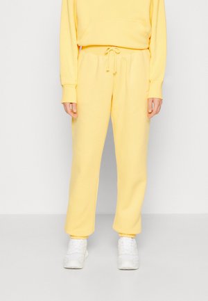 Person iført lysegule træningsbukser med snøre i taljen og matchende sweatshirt, kombineret med hvide sportssko.