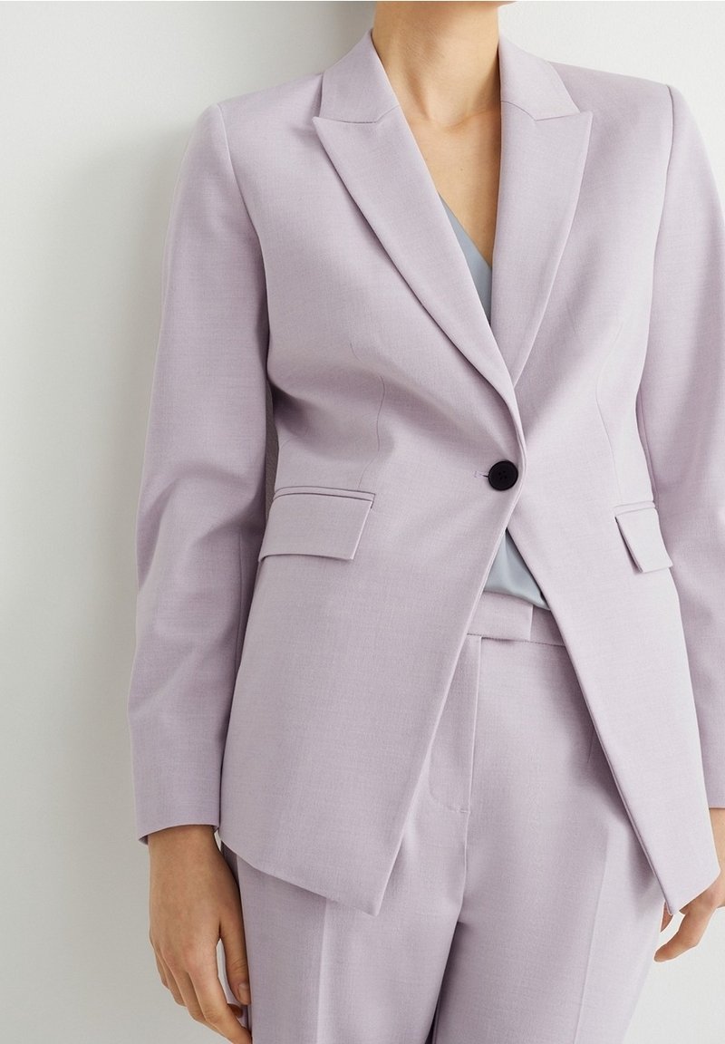 Femme portant un blazer violet clair ajusté avec un bouton noir et un pantalon taille haute assorti, debout contre un mur blanc.