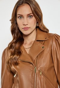 Giacca di pelle marrone con un design aderente, chiusura con zip asimmetrica, dettagli in oro e spalle imbottite. Tessuto liscio.