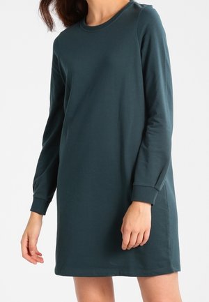 Woman wearing a dark green long-sleeve, knee-length, casual dress with a round neckline and ribbed cuffs.