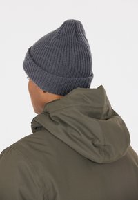 Grijze gebreide beanie met een klep, gecombineerd met een olijfgroene hoodie. Focus op de geribbelde textuur en de relaxte pasvorm van beide items.