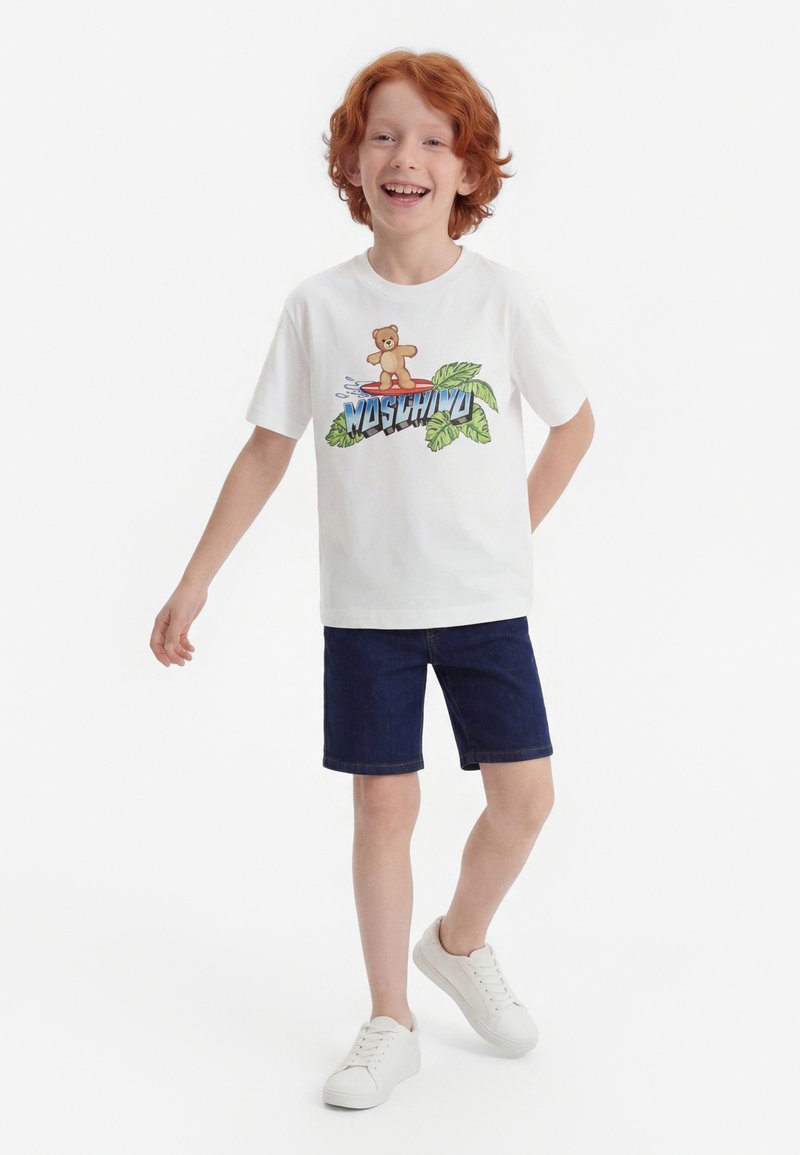 Punapäine poiss naeratab, kandmas valget T-särki, millel on karu pildiga graafika, ja siniseid lühikesi pükse, seistes tavalise valge tausta ees.