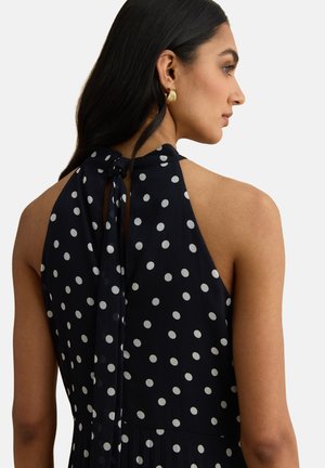 Donna con lunghi capelli scuri che indossa un vestito nero senza maniche con pois bianchi annodato al collo e orecchini a cerchio dorati, di profilo.