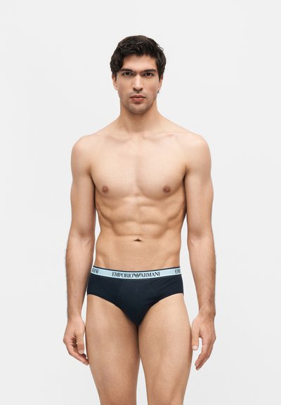 Νεαρός με σκουρόχρωμα μαλλιά που φοράει μαύρα εσώρουχα Emporio Armani, στέκεται μπροστά σε απλό ανοιχτόχρωμο φόντο, κοιτάζοντας ευθεία με ουδέτερη έκφραση.