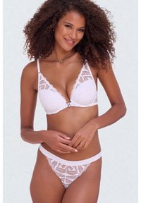 Femme aux cheveux bouclés portant un soutien-gorge en dentelle blanche et une culotte assortie, souriante, les mains délicatement jointes devant un fond clair.