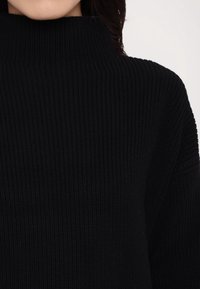 Sort strikket sweater med høj ribstrikket krave, der har en tekstureret overflade og en afslappet pasform, som fremhæver stoffets tykkelse.
