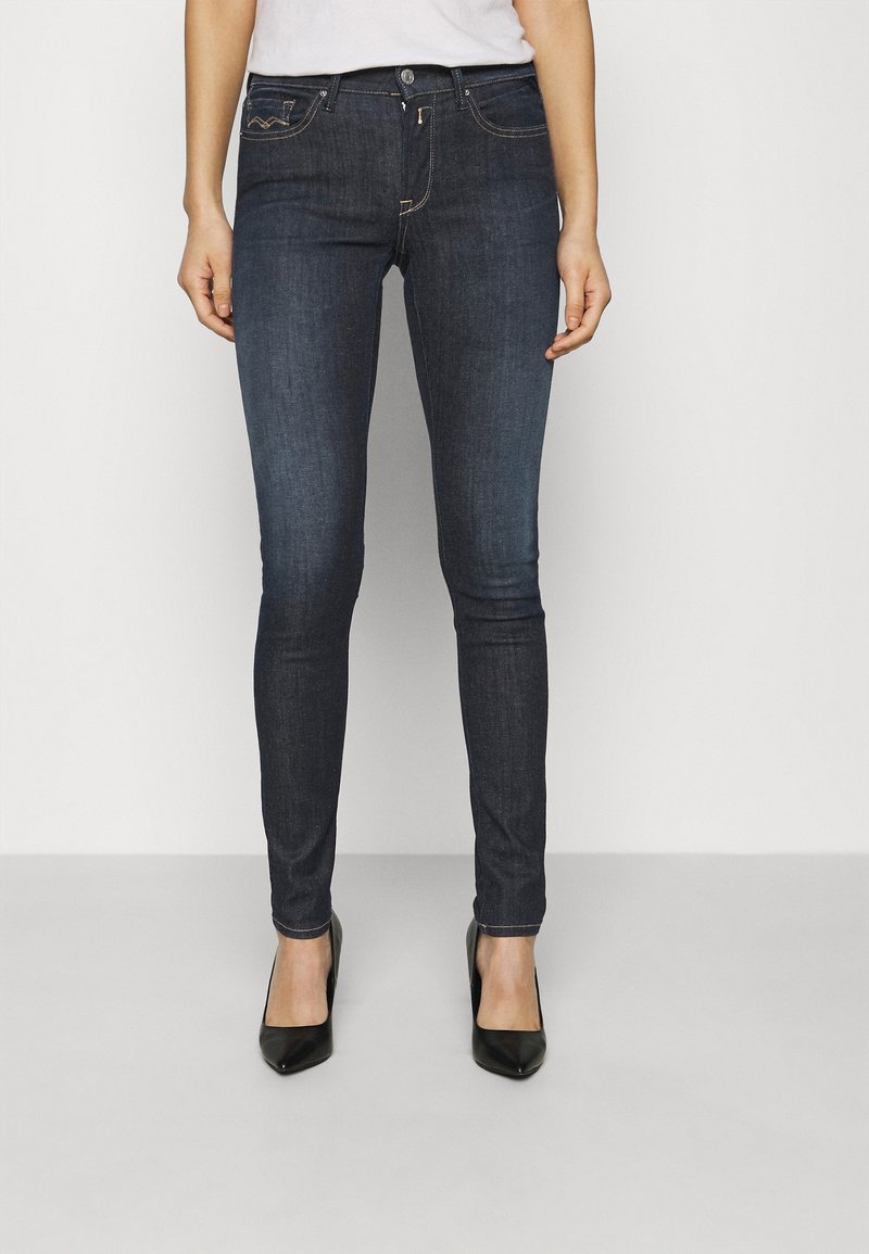 Jeans skinny de denim oscuro con una textura suave, que cuentan con bolsillos frontales y costuras en contraste de color naranja, combinados con tacones negros de punta.