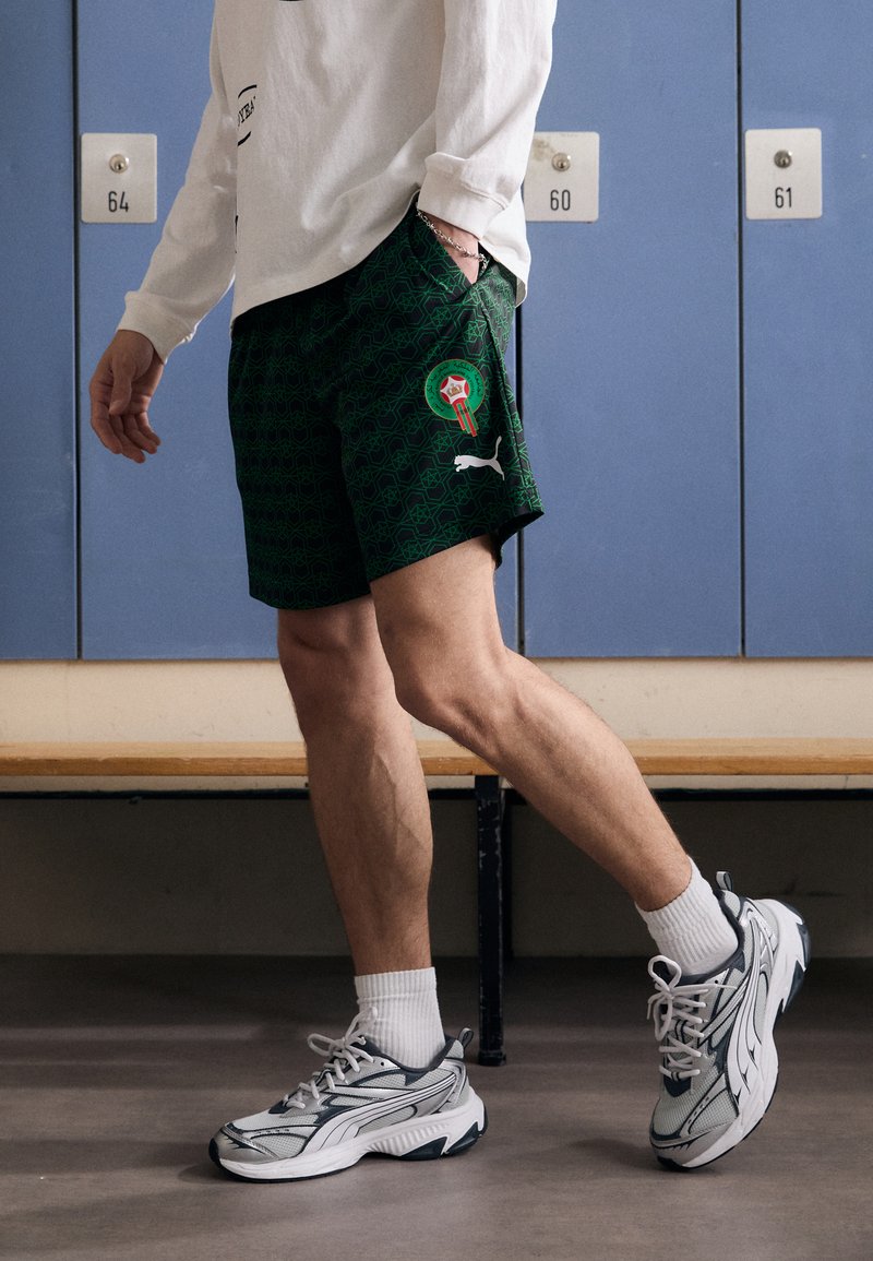 Grønne mønstrede shorts med et marokkansk emblem og hvidt Puma-logo, parret med grå sneakers og hvide crew-sokker.
