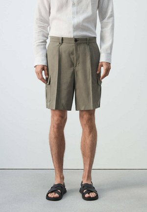 Mann trägt hellbraune Cargo-Shorts, ein weißes Langarm-Hemd mit Knöpfen und schwarze Sandalen mit Kreuzriemen, steht auf einem grauen Boden.