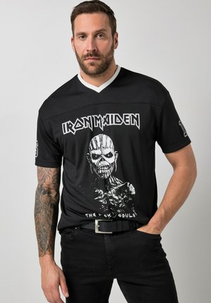 Uomo con barba e braccio tatuato che indossa una maglietta nera della band Iron Maiden con raffigurata una figura scheletrica, in piedi con una mano in tasca.