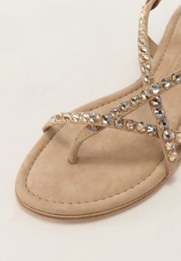 Beige semsket sandal med en tåpost og kryssede stropper prydet med runde, fasetterte klare steiner som dekorasjon.