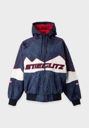 Une personne debout porte une casquette noire, une veste zippée bleu marine et blanche, un sweat-shirt noir à motif, un short en jean bleu clair et des bottes marron foncé jusqu'au milieu du mollet.