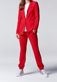 Röd skräddarsydd blazer och jogger-set, tillverkat av ett mjukt tyg. Blazern har notchade slag och fickor; joggarna har elastiska muddar. Kombinerat med vita sneakers.