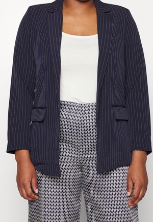 Henkilö, jolla on päällään tummansininen liituraitapuku valkoisen paidan päällä ja korkeavyötäröiset mustavalkoiset kuvioidut housut, seisoo rentoihin asemiin.