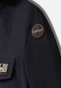 Manche de veste bleu marine foncé avec un écusson rond du logo Napapijri représentant un ours et une bordure orange, et une étiquette en tissu rectangulaire sur la poche.