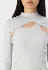 Top lung cu mâneci lungi, de culoare gri deschis, cu textură în relief, având un guler înalt și decupaje pe umeri. Logo mic pe piept.