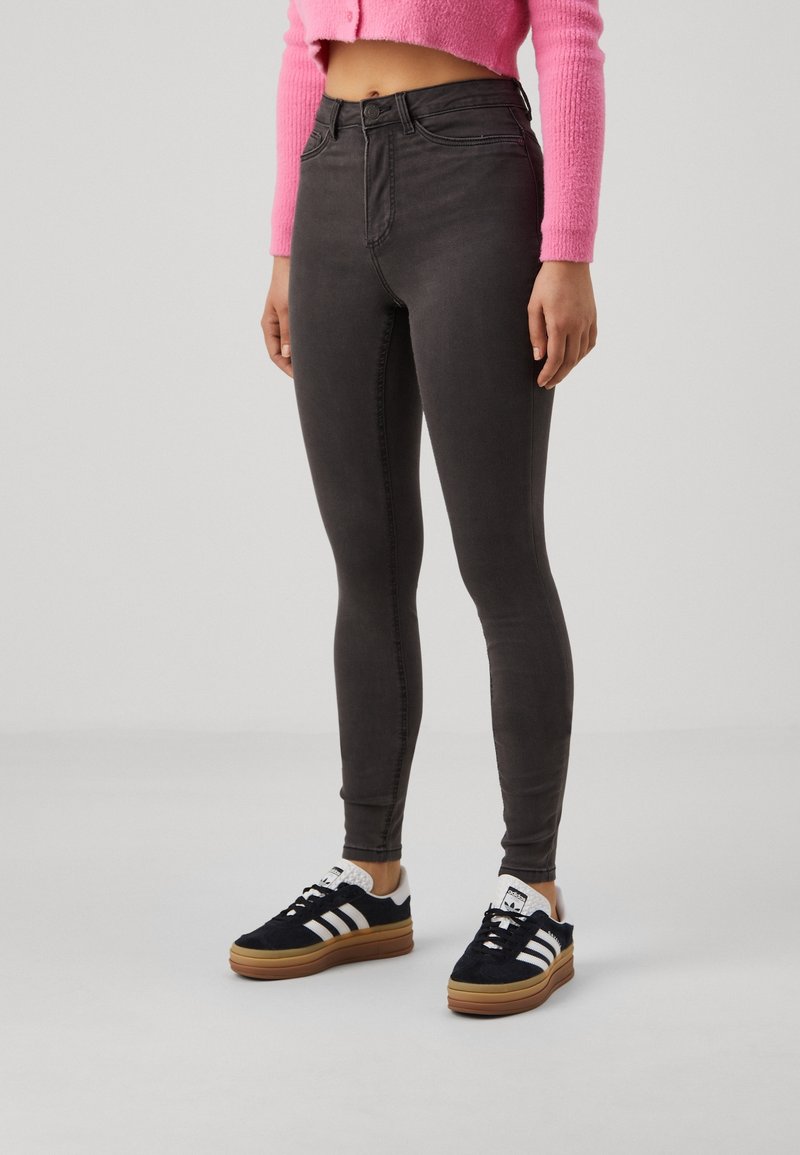 Person som bär högmidjade mörkgrå slim-fit jeans, svarta och vita Adidas-sneakers, samt en kort ljusrosa fluffig tröja.