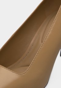 Καφέ δερμάτινο γοβάκι με ψηλό τακούνι, στρογγυλεμένη μύτη και λεπτομερή ραφή κατά μήκος του πάνω άκρου και της εσωτερικής σόλας.