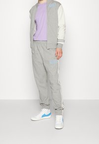 Person som bär en grå Nike träningsdräkt med vita och lavendelfärgade detaljer samt vita sneakers med blå accenter, stående mot en neutral bakgrund.