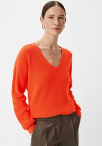 Sweter w serek, oversize, o intensywnym pomarańczowym kolorze, z prążkowaną strukturą i długimi rękawami, w połączeniu z spodniami o wysokim stanie w ciemnym kolorze.