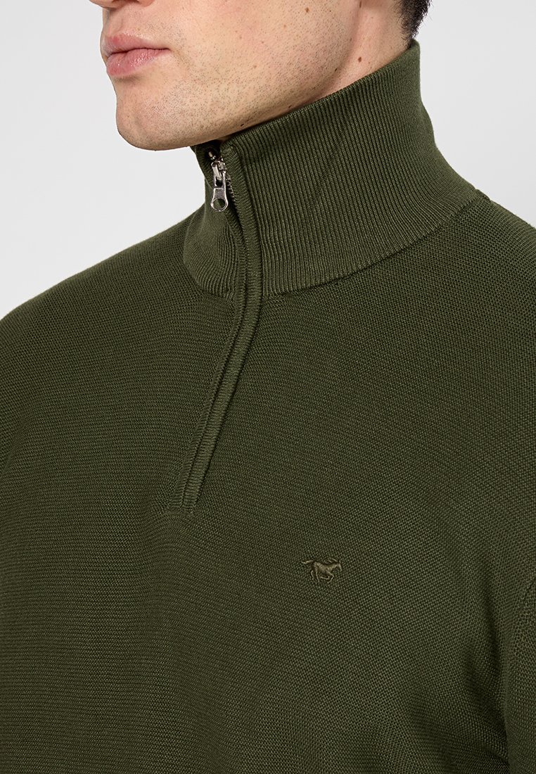 Nahaufnahme eines Mannes, der einen olivgrünen Strickpullover mit einem Viertel-Reißverschluss-Kragen und einem kleinen gestickten Pferdelogo auf der Brust trägt.