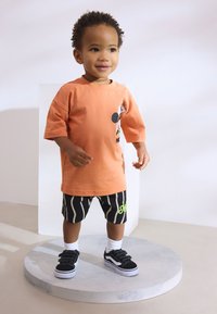 Toddler wearing an orange short-sleeve t-shirt with a graphic, paired with black and white striped shorts, black sneakers, and white socks.