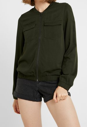 Woman wearing a dark green zip-up jacket with flap pockets and black rolled-up denim shorts, standing with one hand down.