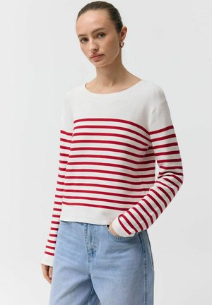 Kobieta ubrana w biały sweter z czerwonymi poziomymi paskami i jasnoniebieskie dżinsy, stojąca z jedną ręką w kieszeni na jednolitym tle.