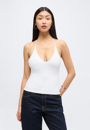 Jeune femme aux longs cheveux raides portant des boucles d'oreilles anneaux, un haut blanc dos nu, un jean foncé et un bracelet en argent, debout devant un fond uni.