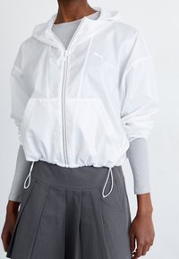 Biała, lekka kurtka z kapturem i elastycznym dołem, zapinana na zamek błyskawiczny z detalem logo. Zestawiona z szarą plisowaną spódnicą.