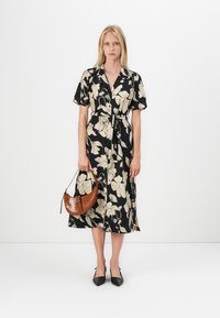 Robe noire à motif floral avec une taille à nouer, des manches courtes et une longueur midi, associée à un sac à main texturé marron et des escarpins noirs à bout pointu.